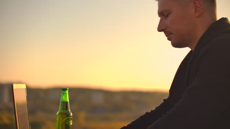 A-male-freelance-programmer-sits-on-a-skyscraper-roof-with-a-laptop-and-beer-typing-code-on-a-keyboard-during-sunset.-Remote-work